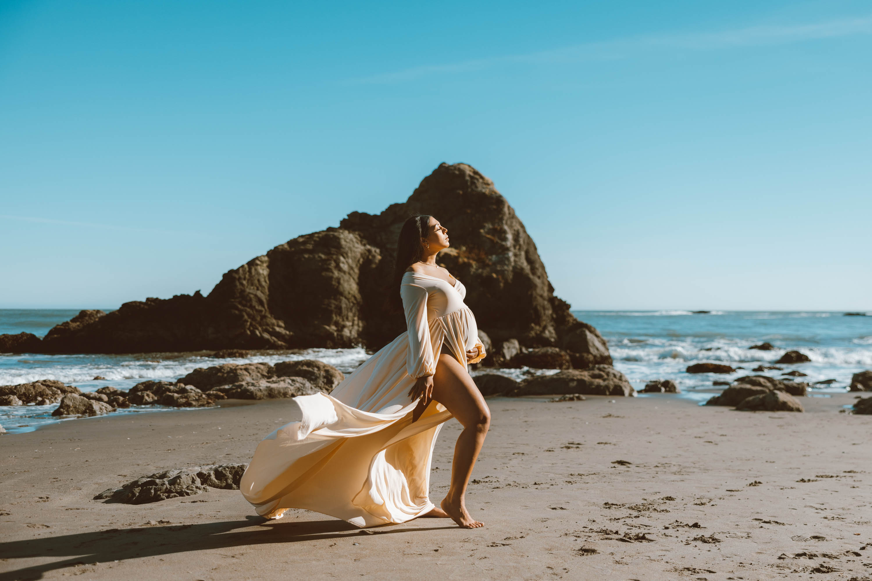 Black mother to be standing on beach in front of rock with beige golden dress during maternity photo session