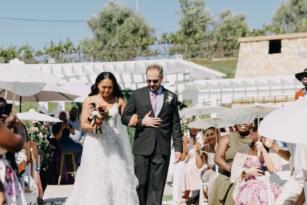 Black bride walking down the aisle at an outdoor vineyard wedding ceremony, guests standing on either side.