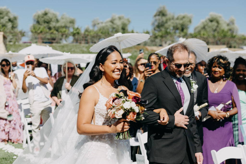 Black bride walking down the aisle at an outdoor vineyard wedding ceremony, guests standing on either side.