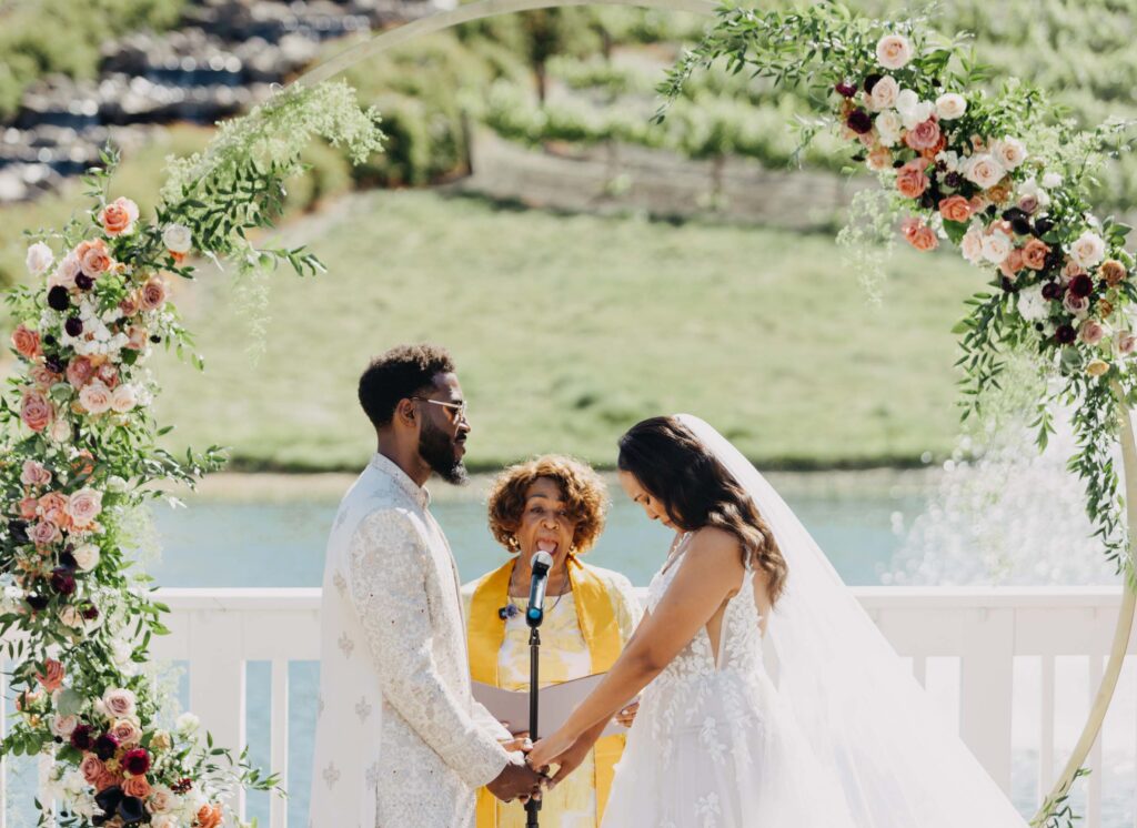 Black bride and groom exchanging vows under a floral arch at Lavanda Leal Vineyards, rows of grapevines in the background.