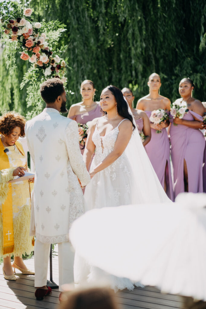 Black bride and groom exchanging vows under a floral arch at Lavanda Leal Vineyards, rows of grapevines in the background.
