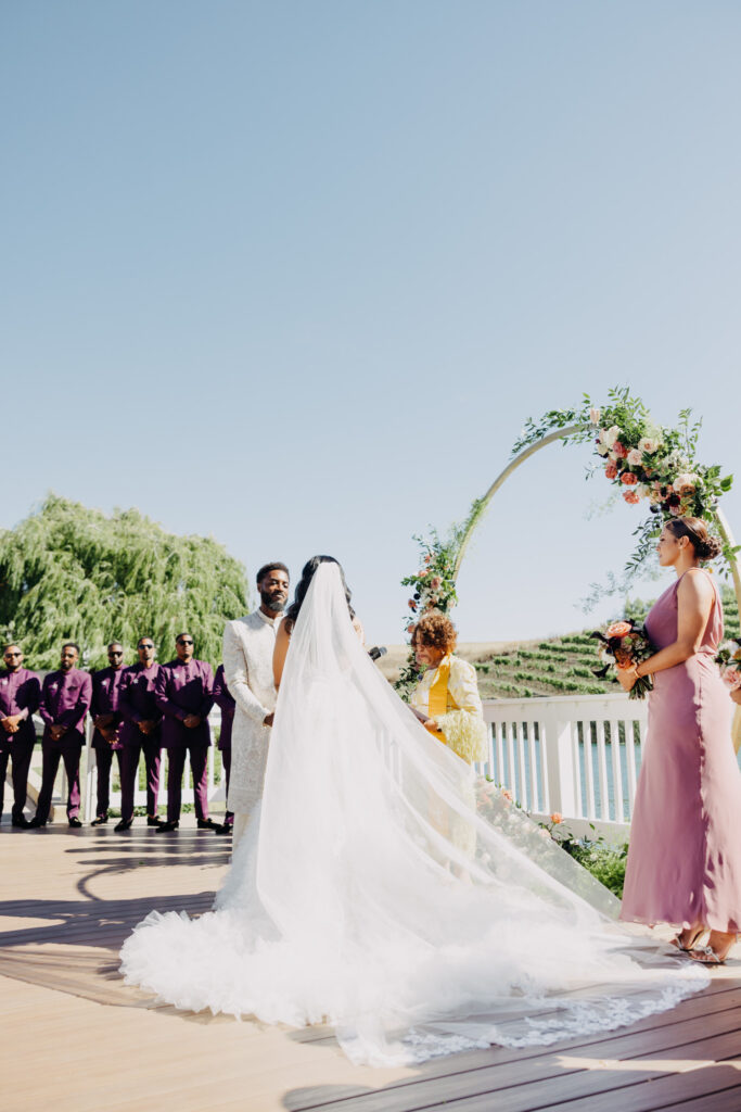 Black bride and groom exchanging vows under a floral arch at Lavanda Leal Vineyards, rows of grapevines in the background.