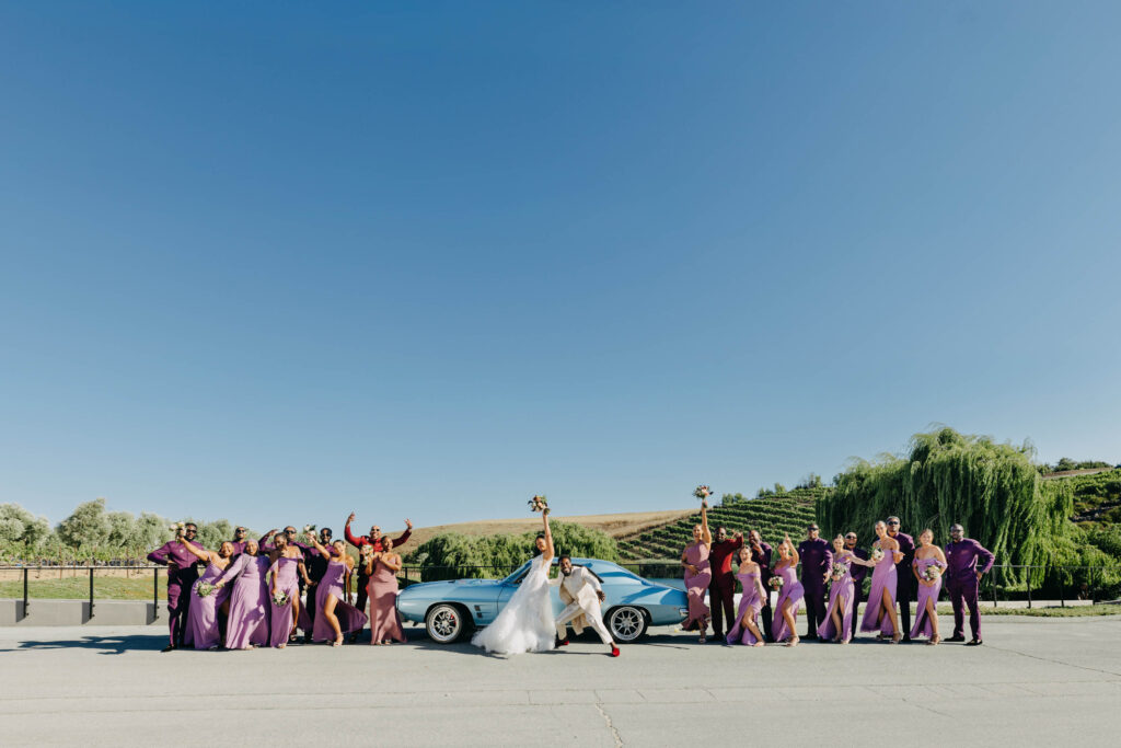 Black bride and groom having fun with their wedding party in front of vintage blue car at golden hour at Lavanda Leal Vineyards.